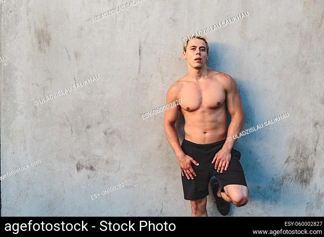 Natural portrait of young athletic shirtless man standing near concrete wall