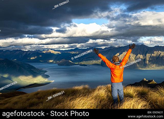 Hiker stretches his arms in the air, view of Lake Hawea in the evening light, lake and mountain landscape, view from Isthmus Peak, Wanaka, Otago, South Island