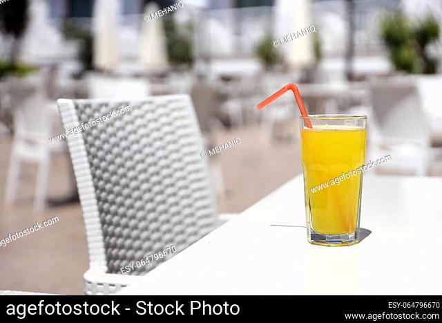 Glass of orange juice with plastic straw on white table in restaurant outdoor lounge zone in sunny weather