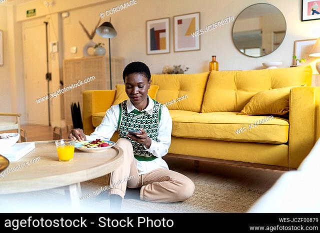 Woman using mobile phone and eating breakfast in living room