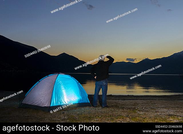 Man and an Illuminated Tent on Alpine Lake Maggiore with Mountain in Dusk in Ascona, Switzerland