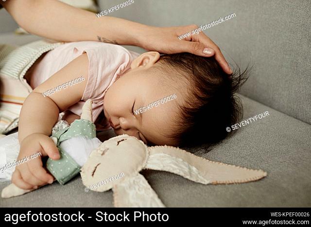 Mother stroking daughter sleeping with stuffed toy on sofa at home