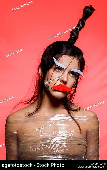 Beautiful young woman with odd fancy hairstyle and eyebrows and lips paper cutouts on face. Beauty shot on red background. Copy space