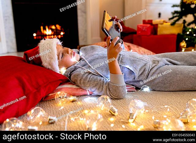 Side view of a happy young Caucasian boy lying on the floor, wearing Santa hat and a onesie, using a tablet in his sitting room at Christmas time