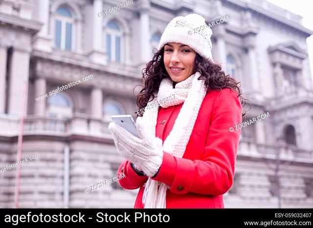 Beautiful Woman in Red Coat and and wool cap and gloves read messages on smartphone and smiling. Urban Space. Cold Weather