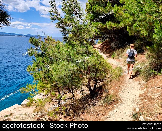 Young active feamle tourist wearing small backpack walking on coastal path among pine trees looking for remote cove to swim alone in peace on seaside in Croatia