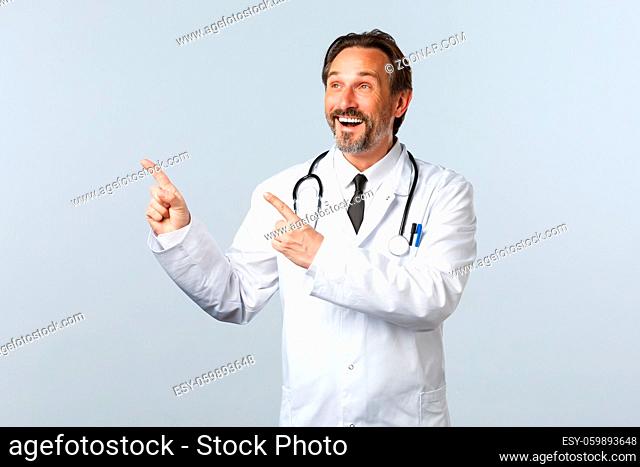 Covid-19, coronavirus outbreak, healthcare workers and pandemic concept. Happy smiling male doctor in white coat pointing fingers upper left corner amused