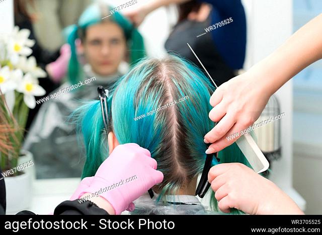 Back view of female head with emerald hair color and regrown hair roots. Woman sits in chair by mirror, two professional hairdressers combing client's hair...
