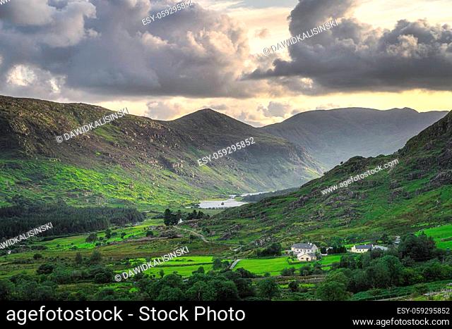 Small cottage surrounded by forest, lake and river in Black Valley. MacGillycuddys Reeks mountains illuminated by sunlight, Ring of Kerry, Ireland