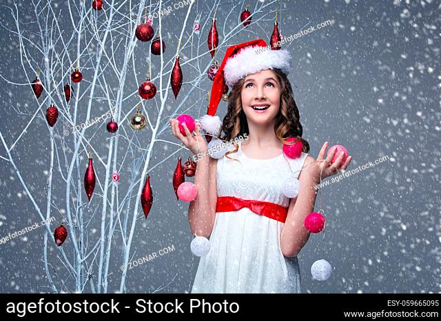 Beautiful teen girl with long curly hair in white dress with red ribbon belt and santa cap on head standing near white tree branches wiht christmas decorations...
