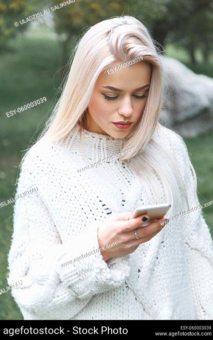 Portrait of young caucasian blonde hair woman in white pullover uses the phone outdoor