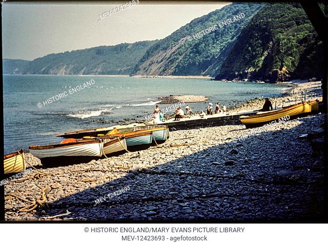 A view looking south-east across the foreshore of Clovelly Harbour, with small boats beached on the shingle and people sitting and standing on the slipway and...