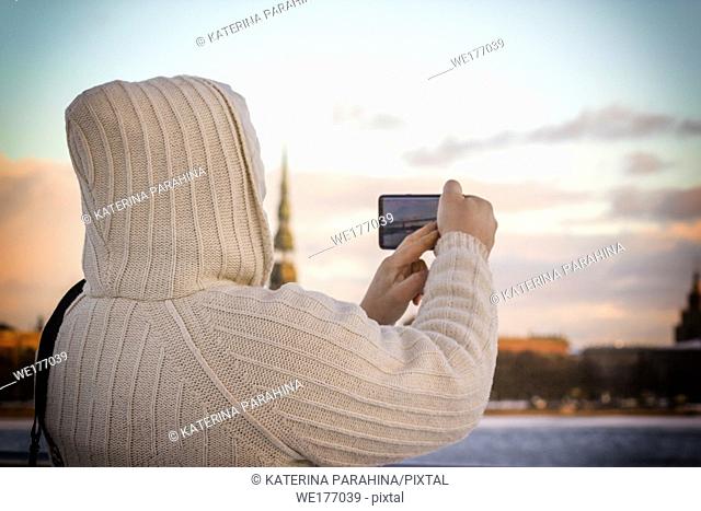 Man in white knitted jacket taking photo of cityscape with his smartphone in Riga, Latvia