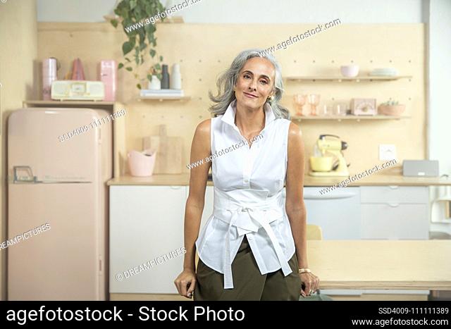 Wide horizontal portrait of a middle-aged woman with gray hair, standing in a kitchen looking into camera with sincere smile