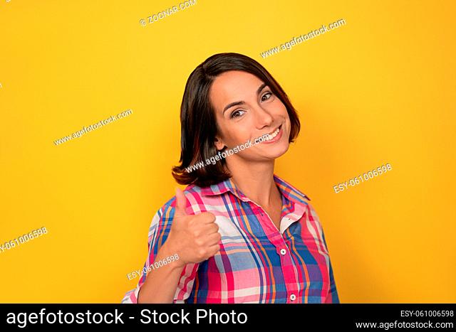 Raising thumb up gesture with hand smile beautiful woman dressed in a plaid shirt and dark hair on yellow background. Human emotions, facial expression concept