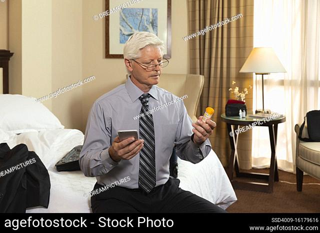 Mature Caucasian man sitting on edge of bed in a hotel room, using cell phone to check prescription medication