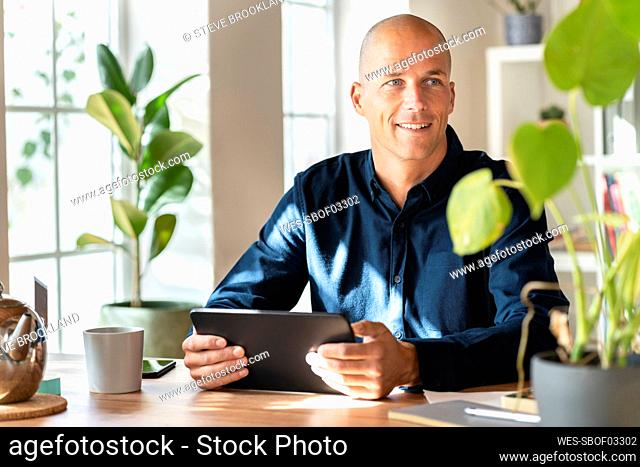 Smiling businessman holding digital tablet while looking away in home office