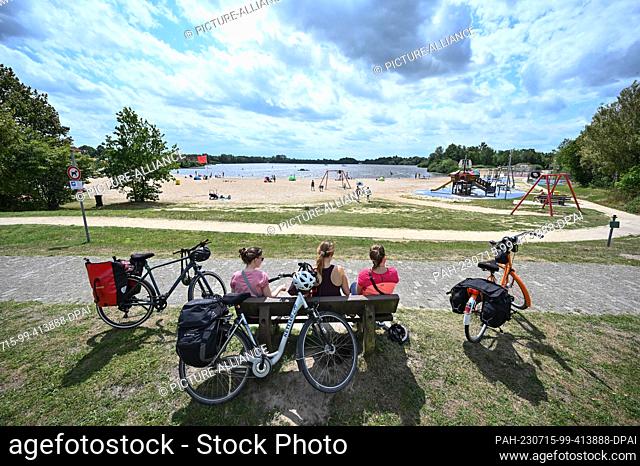 15 July 2023, Timmel: Bicycle tourists take a short break at the bathing lake and enjoy the weather. Weekend vacationers and tourists have sought cooling on the...