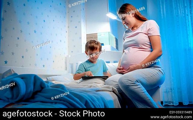 Young pregnant woman sitting at her little son bed and looking at him playing games on tablet computer. Children and adults using smartphones and gadgets at...