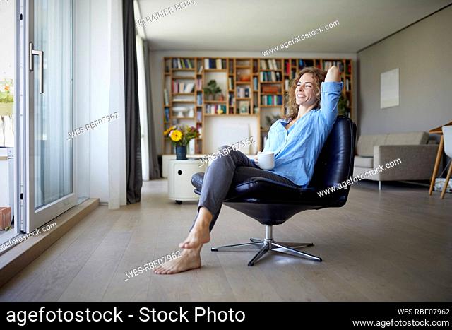 Smiling woman with hand behind head drinking coffee while sitting on chair at home