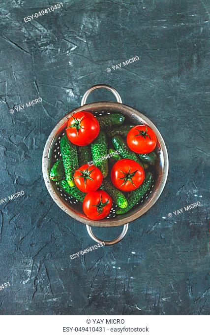 raw whole washed tomatoes and cucumbers in a colander on a dark concrete background. View from above. Flat lay