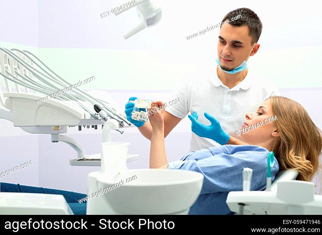 Female patient in blue shirt on the patient chair in the dental cabinet. Next to her there is a male dentist in a white uniform with blue latex gloves and a...