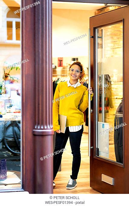 Young woman standing in door of a fashion store, holding laptop