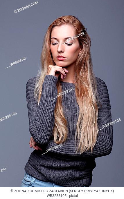 Portrait of young woman in gray woolen sweater and jeans. Beautiful girl posing on grey studio background. Female with blonde hair and day beauty makeup