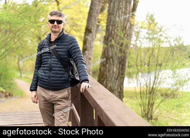 A middle-aged man in sunglasses and a thin down jacket on a wooden footbridge near the lake in early spring