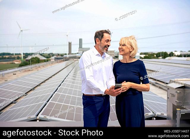 Happy businessman sharing mobile phone with businesswoman in front of solar panels on rooftop