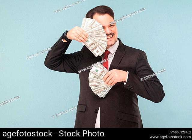 Businessman hides behind fans out of money. Indoor, studio shot, isolated on light blue or gray background