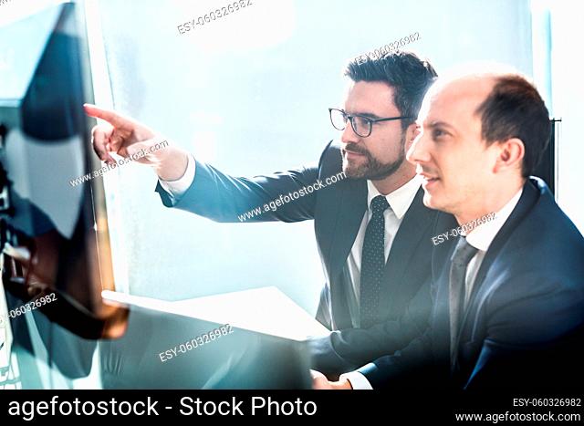 Image of two thoughtful businessmen looking at data on multiple computer screens, solving business issue at business meeting in modern corporate office