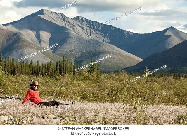 Young woman relaxing, enjoying evening light, sitting in the grass, Cotton Grass, Peel Watershed, Northern Mackenzie Mountains behind, Wind River