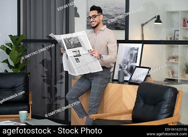 Morning news. Cheerful young bearded man in glasses standing reading interested newspaper at home in morning
