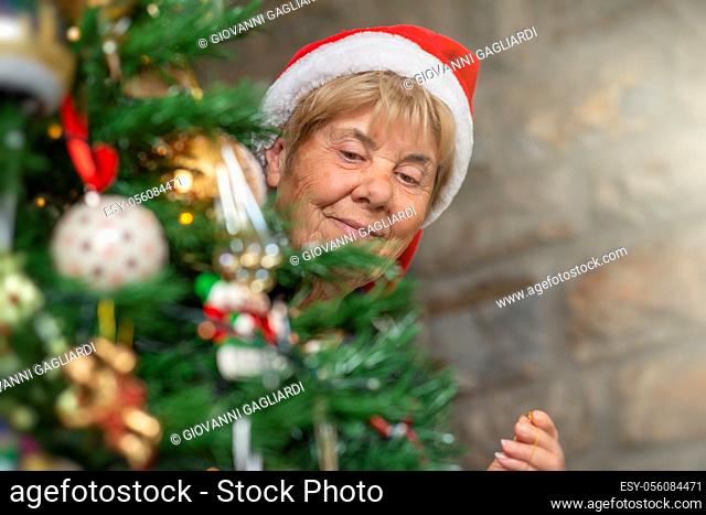 Happy elderly retired woman wearing Santa Red Hat decorating Christmas Tree at home