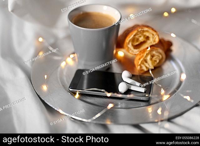 smartphone, earphones, coffee and croissant in bed