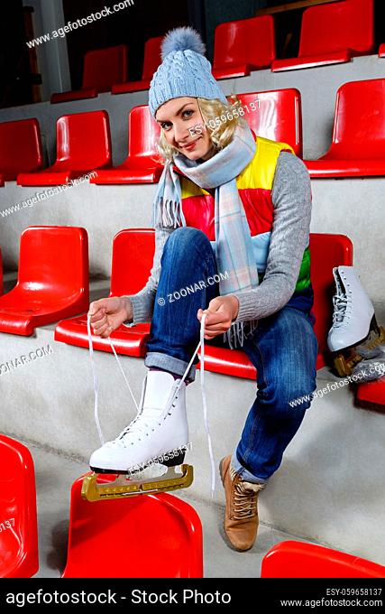 Beautiful blond young woman in winter clothes sitting on red tribune and putting on white skates. Girl getting ready for skating on ice rink