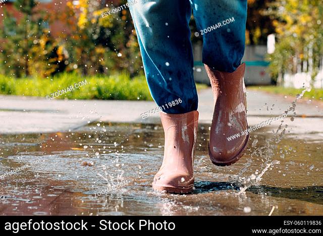 Woman wearing rain rubber boots walking running and jumping into puddle with water splash and drops in autumn fall