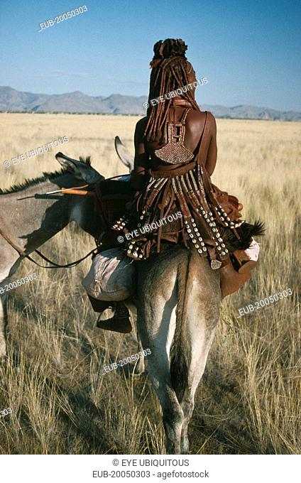 Himba tribeswoman on donkey seen from behind wearing typical leatherwork decorated with cowrie shells and copper