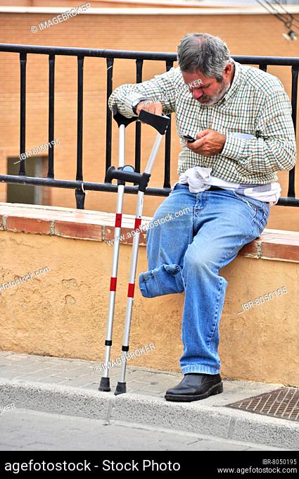 One-legged man with crutch sits on wall and looks into his mobile phone, Villaricos, Andalusia, Spain, Europe