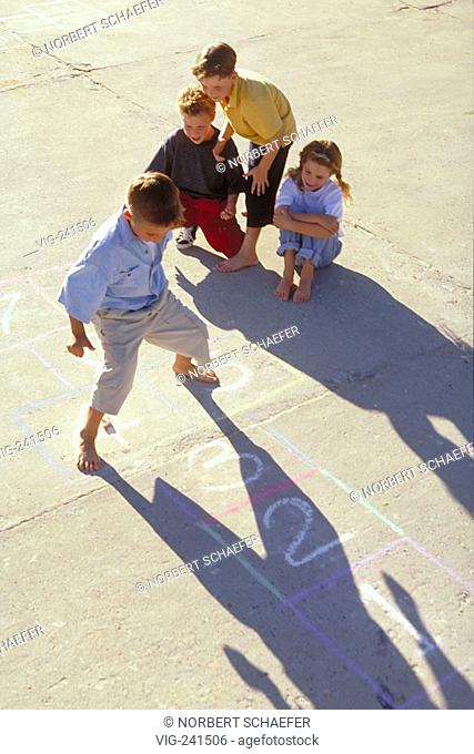 portrait, long shadows of a group of 3 children jumping bare-feeted on squares with numbers drawn with chalk on the asphalt  - GERMANY, 05/04/2004