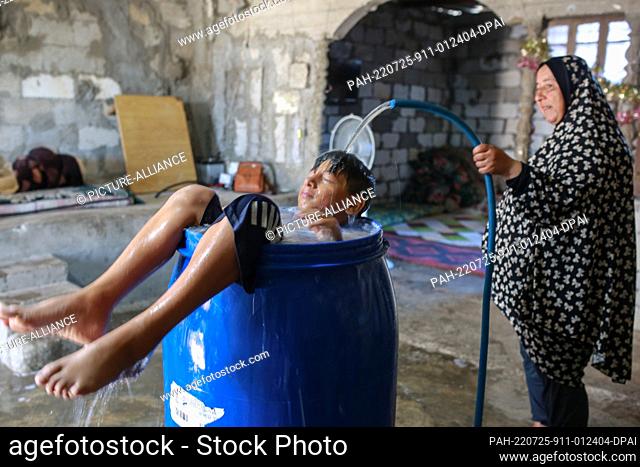 25 July 2022, Palestinian Territories, Khan Yunis city: A Palestinian child takes a bath in a plastic bowl, inside his sheet metal home