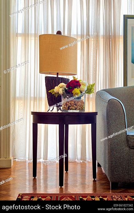 Table lamp and colorful flowers planter on small dark brown wooden table on background of big window with white sheer curtains and grey armchair