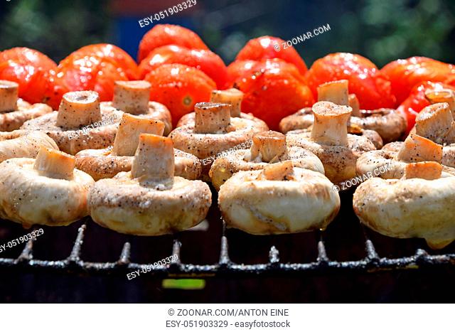 Vegetables in salt and spices being cooked on char grill outside, white champignons portobello mushrooms and red small tomatoes