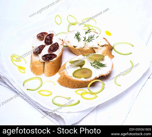 sandwiches with creamy white cheese, sausage, olives and dill on a white plate, wooden white table
