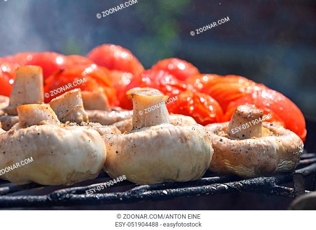 Vegetables in salt and spices being cooked on char grill outside, white champignons portobello mushrooms and red small tomatoes, close up, low angle view