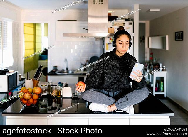 Teenage girl with apple fruit listening music through headphones while using mobile phone at home