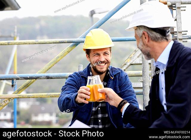 Happy architect and worker on scaffolding on a construction site clinking juice bottles