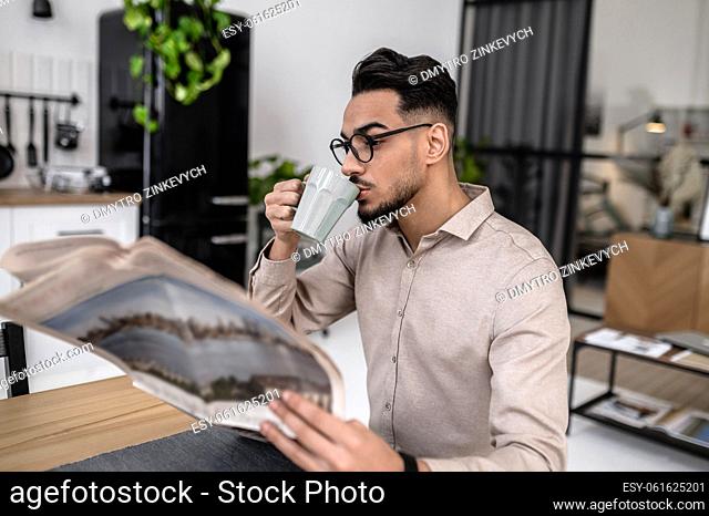 Interesting news. Young bearded engaged man drinking coffee reading newspaper sitting at table sideways to camera in modern apartment