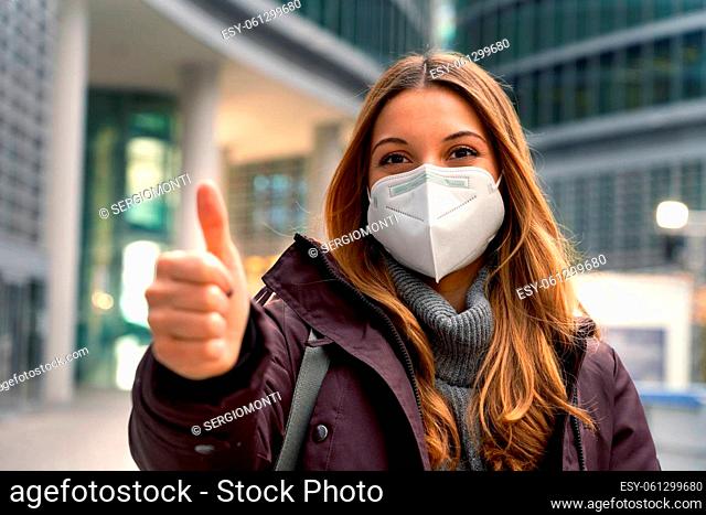 Close up of beautiful confident woman wearing protective mask KN95 FFP2 showing thumb up with modern city on background and looking at camera